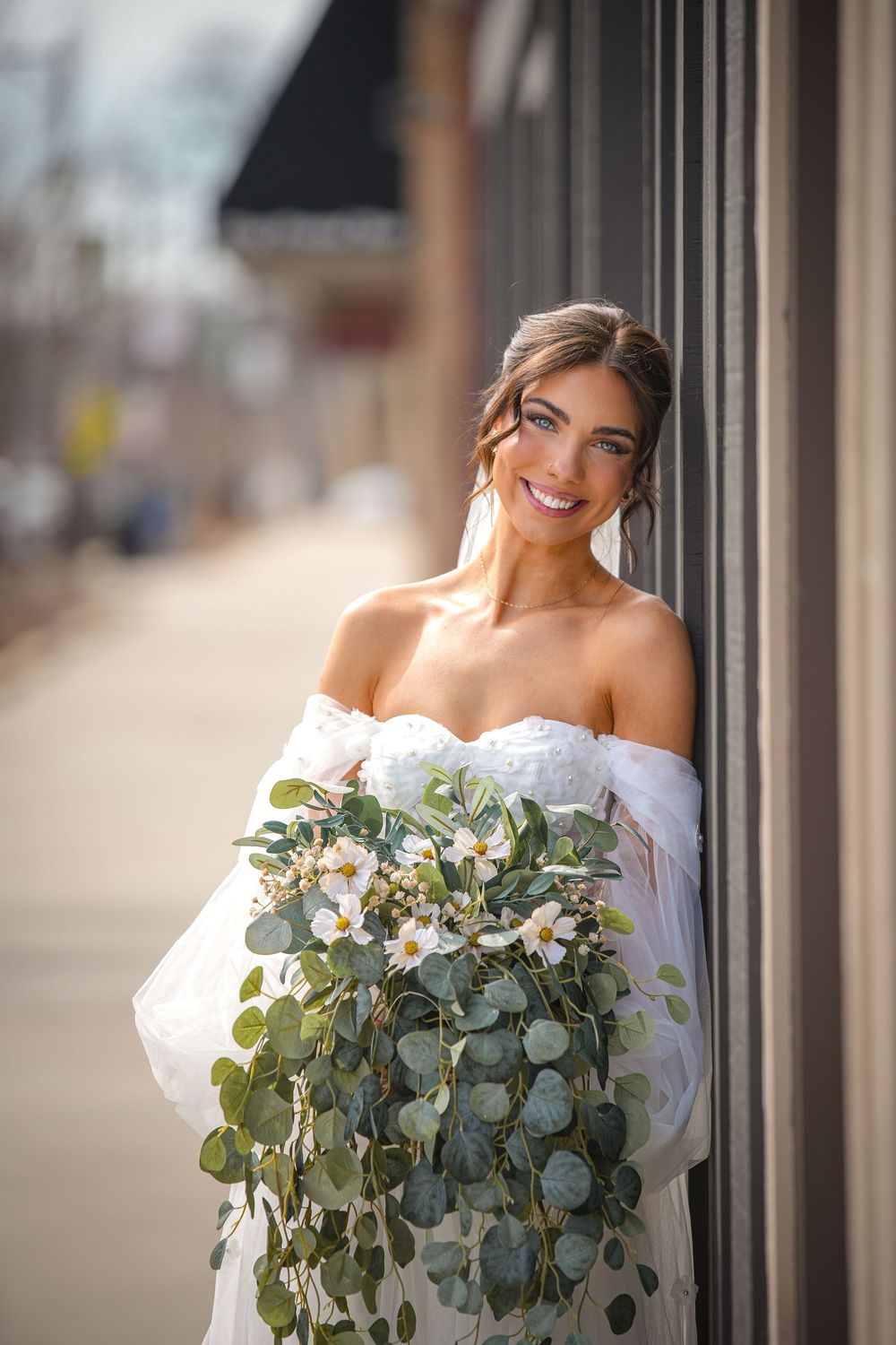Smiling bride in off-shoulder white gown holds cascading eucalyptus and white flower bouquet outdoors.