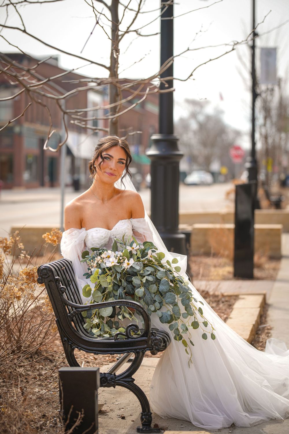 Elegant bride in off-shoulder white gown sits on park bench holding cascading eucalyptus bouquet on city sidewalk.