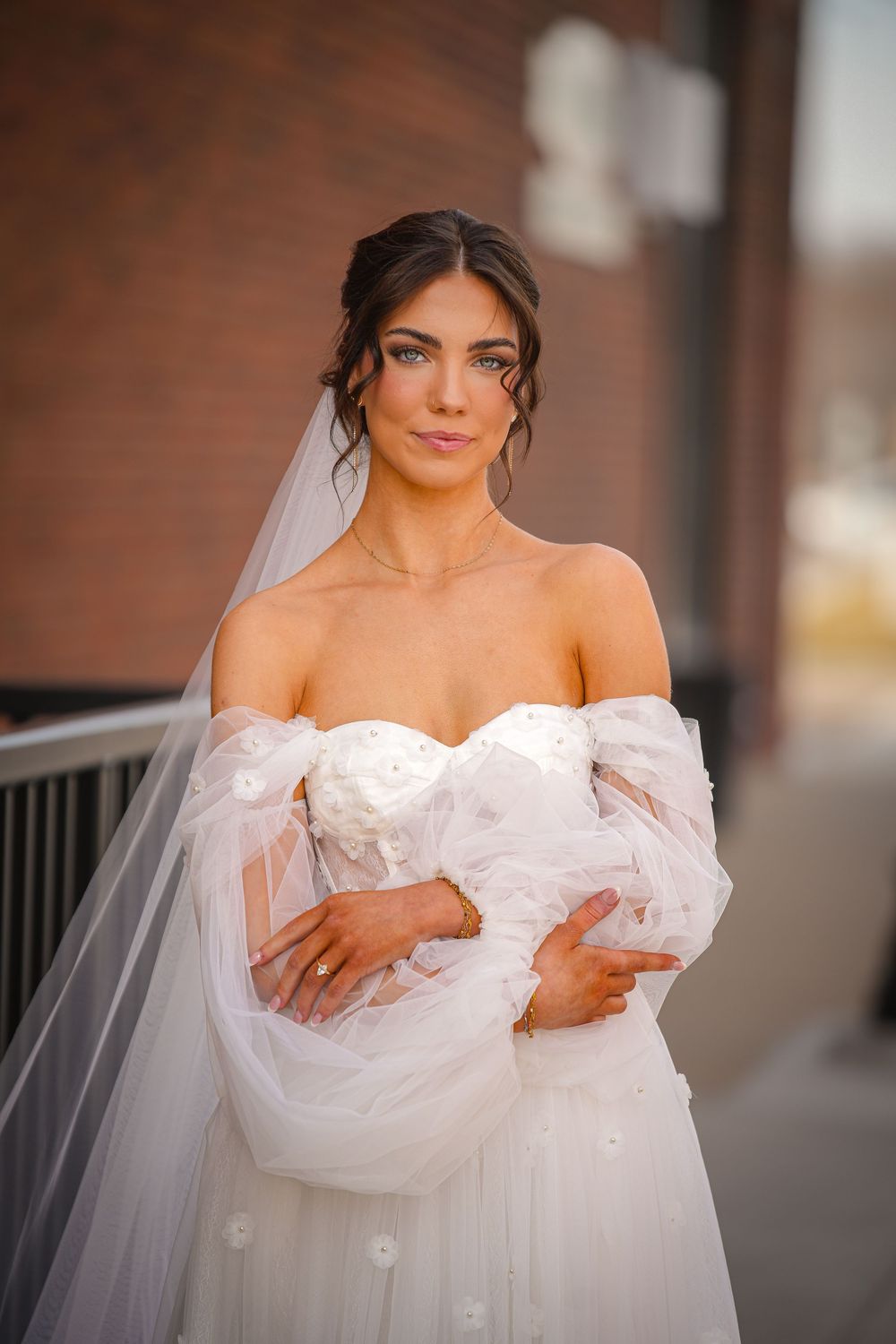 Elegant bride in off-shoulder white tulle gown with floral details and veil, posing outdoors near brick building.