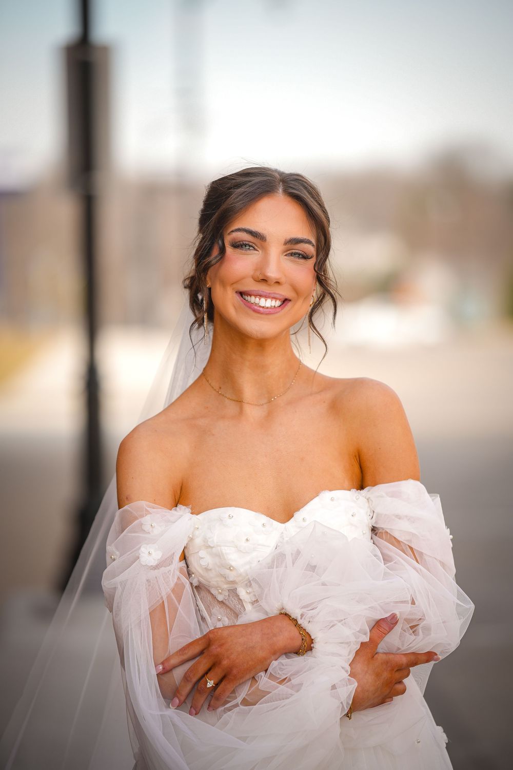 Radiant bride in off-shoulder white tulle gown with veil smiling outdoors on wedding day.
