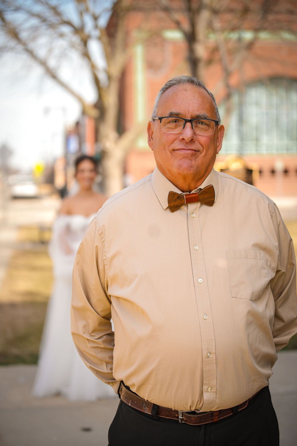 Older man in tan shirt and brown bow tie smiles outdoors with bride visible in background.