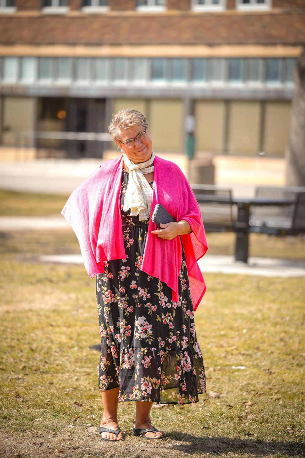 Older woman in black floral dress and pink shawl holds book while standing outdoors in sunlight.