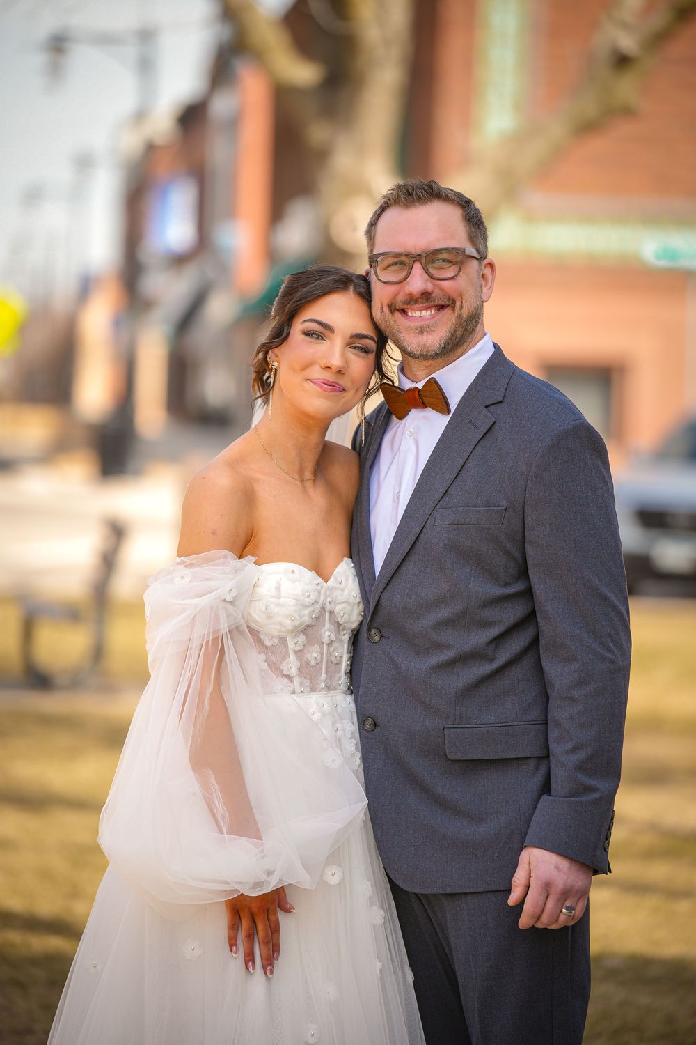 Smiling bride and groom pose outdoors in urban setting, she in white gown, he in gray suit with bow tie.