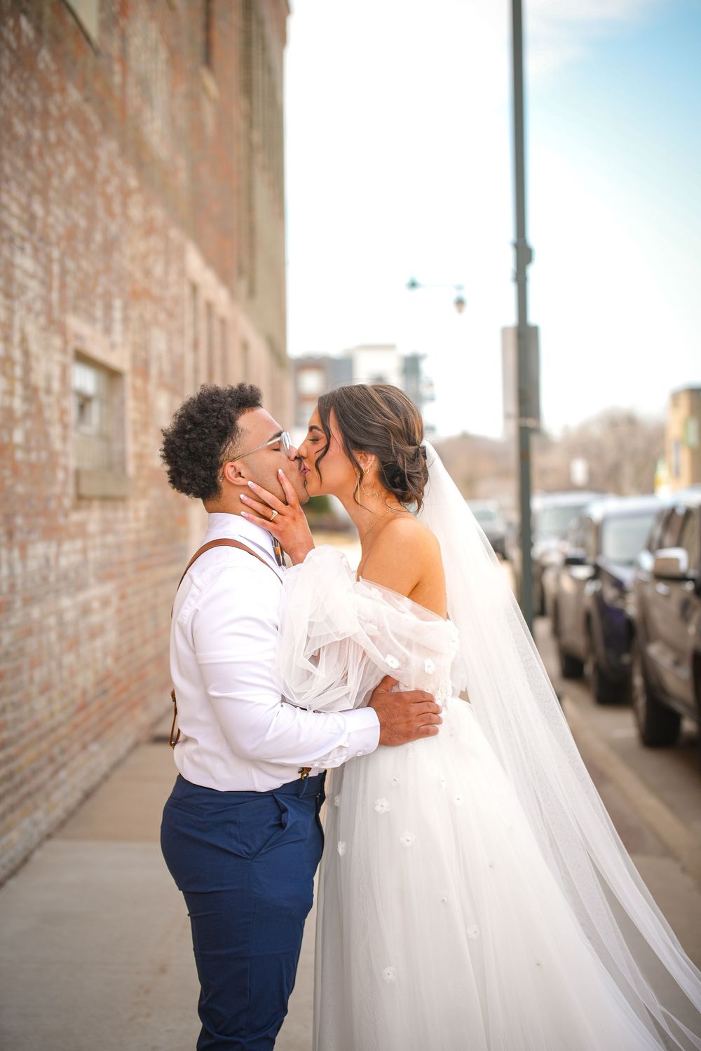 Bride and groom share romantic kiss on urban sidewalk, bride in off-shoulder gown with veil.