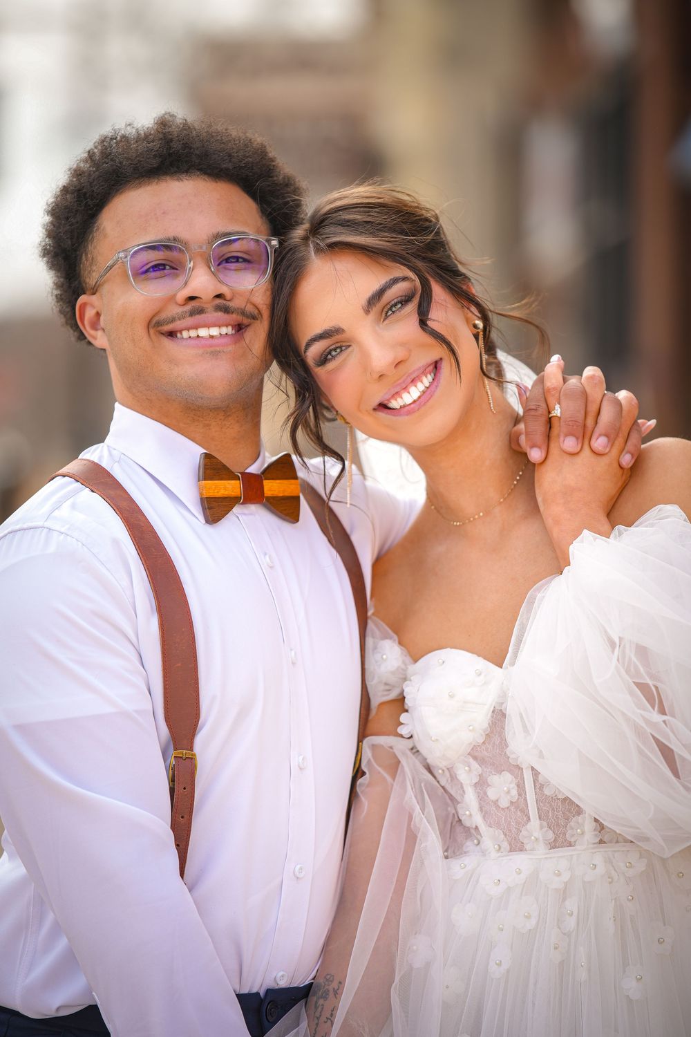 Joyful interracial couple in wedding attire smile together, groom in suspenders and bow tie outdoors.