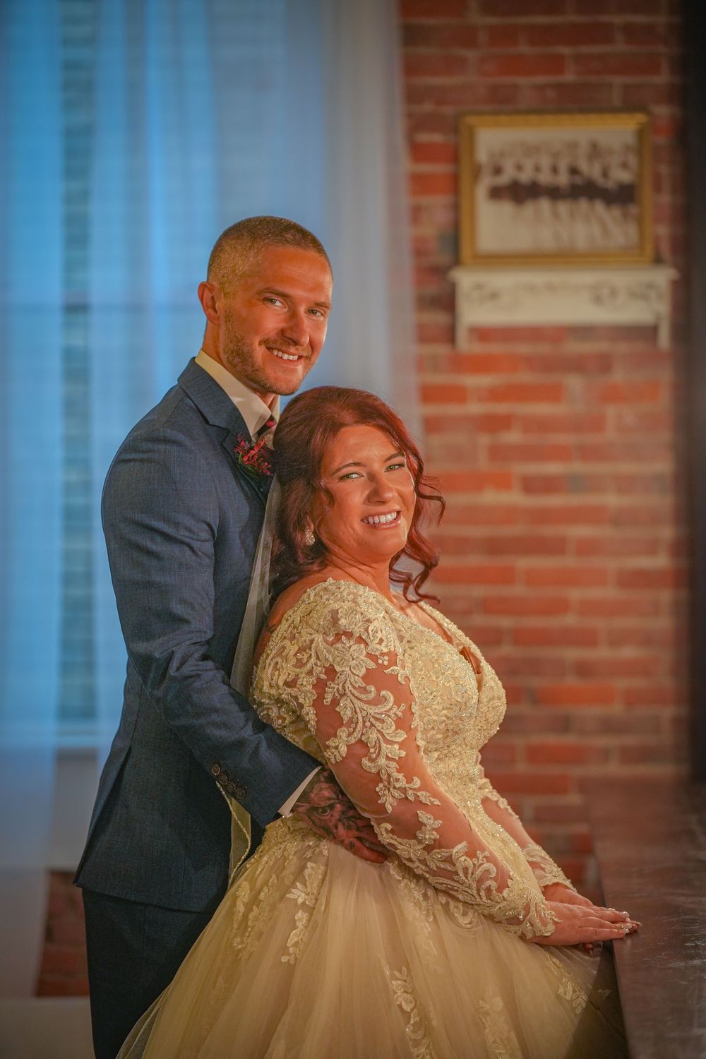 A joyful wedding couple embraces in front of a rustic brick wall with soft window lighting.