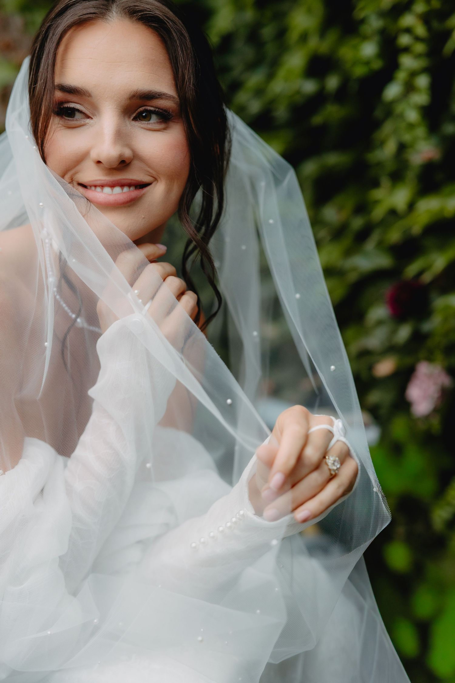 Smiling bride in a sheer pearl-dotted veil and white lace sleeves adjusts her veil, showing her ring against a lush green ivy Utah backdrop.