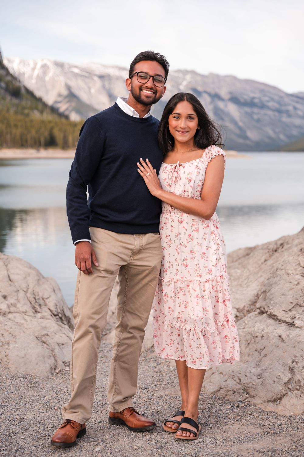 A couple poses together near a mountain lake, wearing casual dressy attire in a scenic outdoor setting.