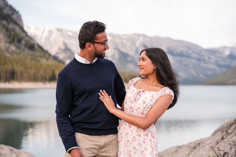 A couple stands close together at the edge of a mountain lake with snow-capped peaks in the background.