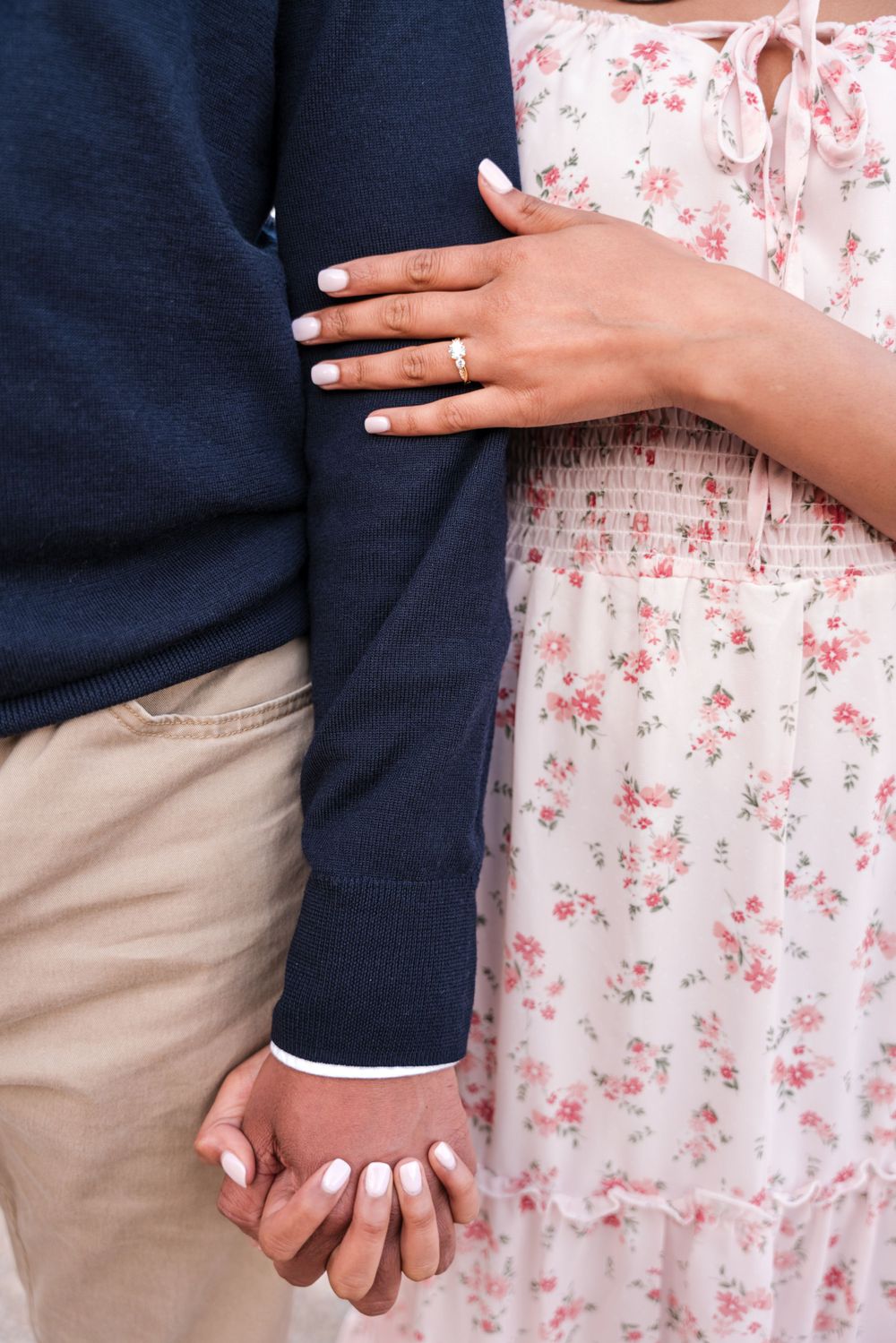 Close-up of entwined hands showing an engagement ring against floral dress and navy sweater.