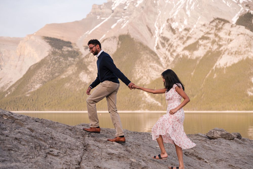 Two people walk along a rocky ridge with majestic mountain peaks and a lake in the scenic wilderness backdrop.