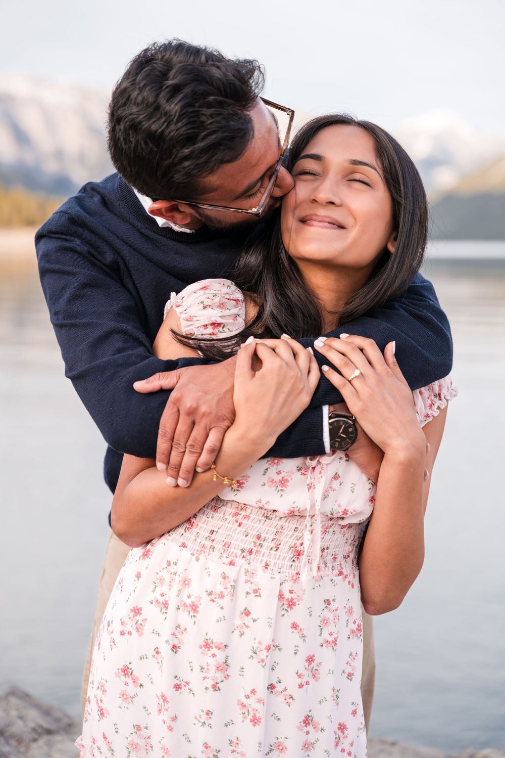 A romantic couple embraces by a mountain lake, with one wearing a floral dress and the other in a navy sweater.