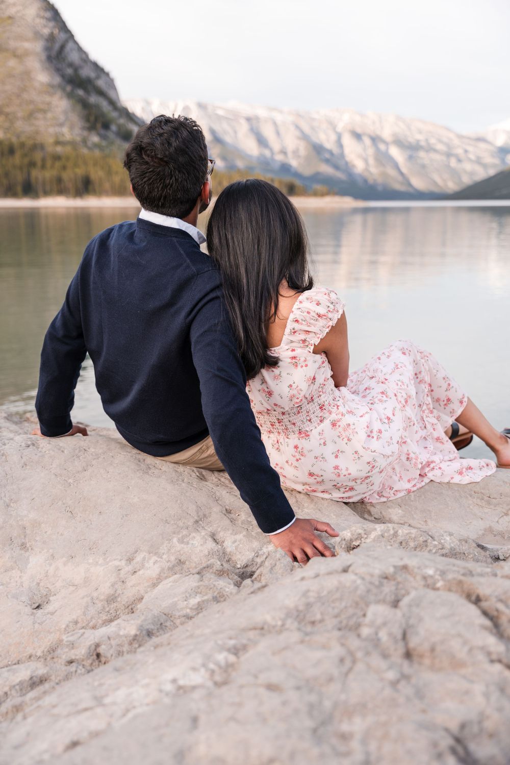 A couple sits together on rocky terrain overlooking a mountain lake at sunset.