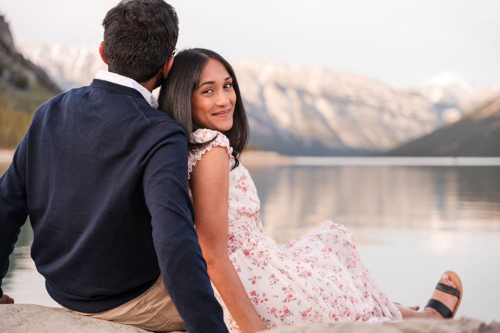 A couple sits together on a dock by a scenic mountain lake at sunset while wearing formal attire.