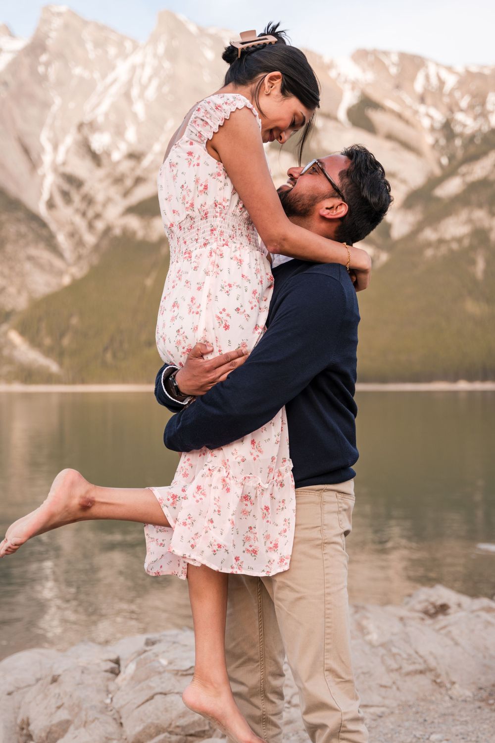 A romantic couple embraces by a mountain lake, with her wearing a floral dress and him in a navy sweater.