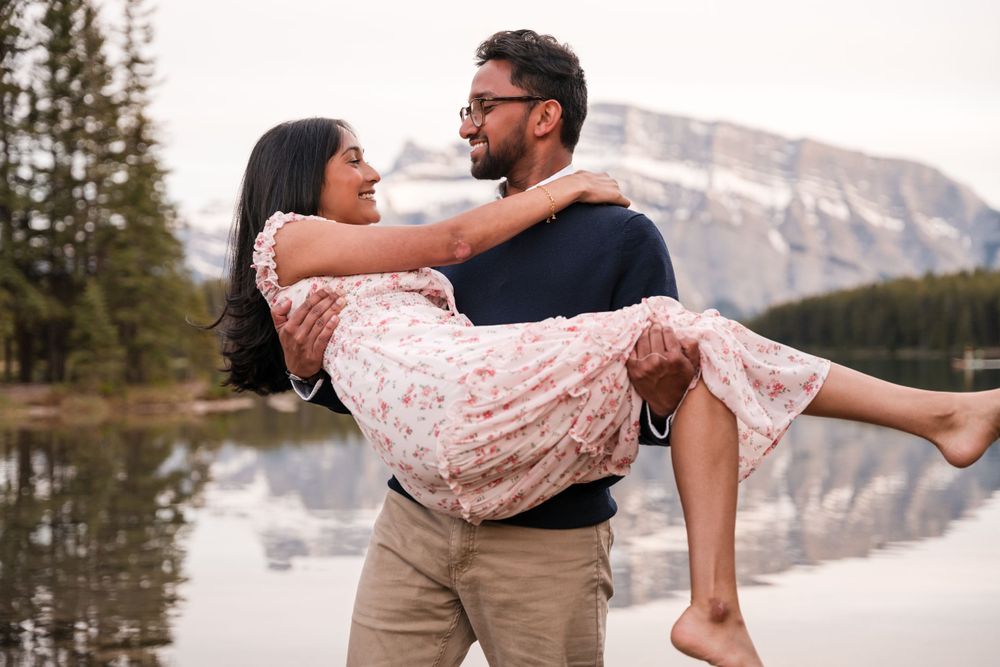 A romantic couple shares a playful moment as one carries another by a mountain lake at sunset.