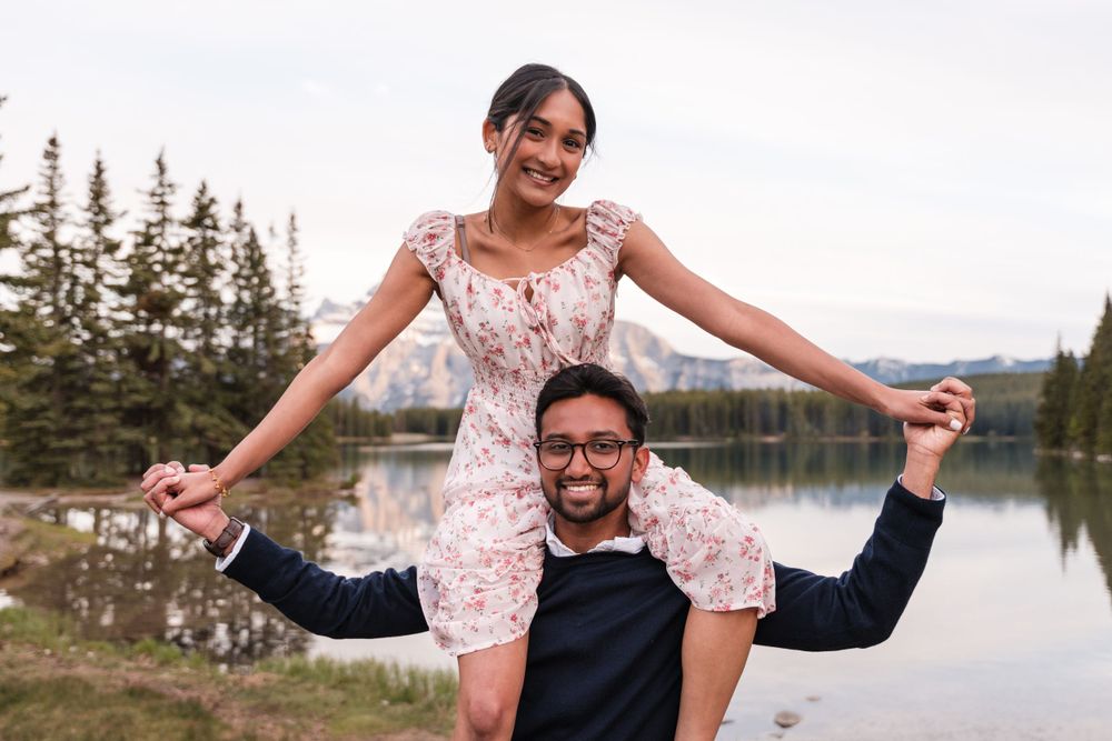 A couple poses joyfully by a serene lake with pine trees in the background during golden hour.