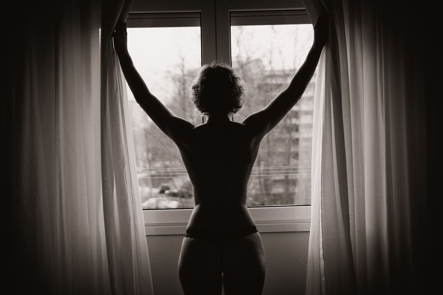 Silhouette of person stretching arms up by window with curtains.
