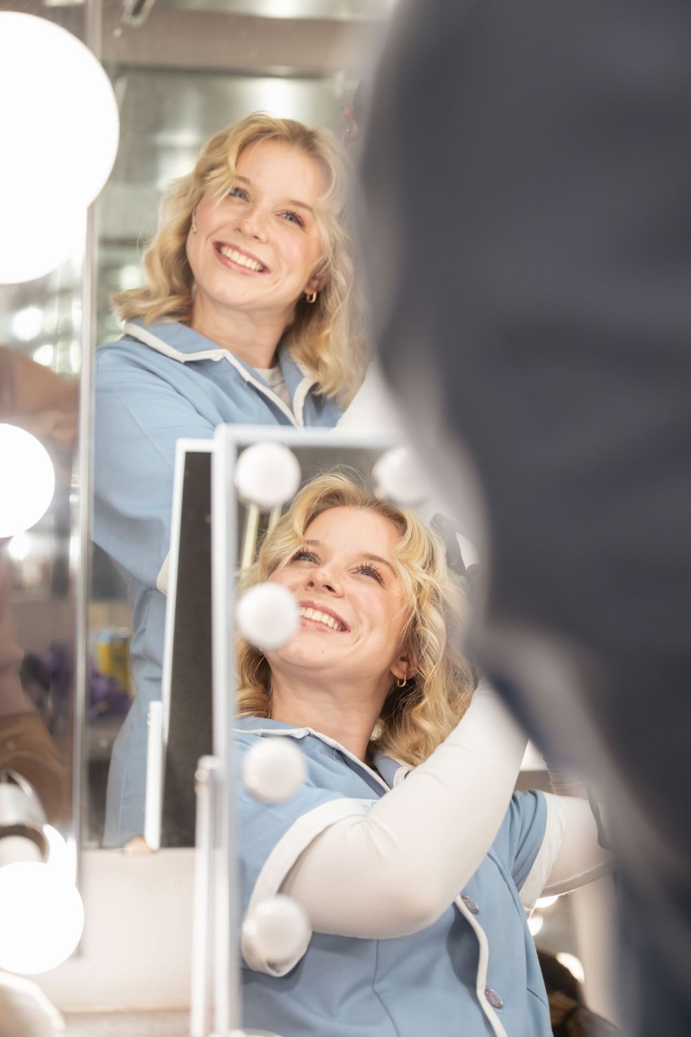 A reflection of someone in a blue top smiling brightly in a vanity mirror with illuminated bulbs.