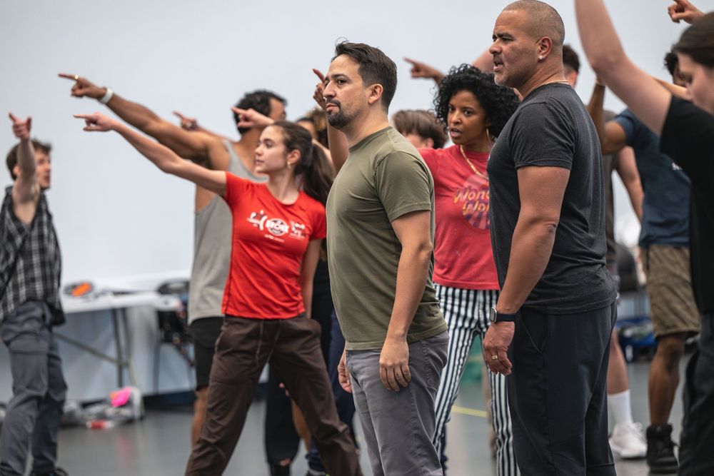 A group of performers rehearsing choreography together on a stage with their arms extended outward in unison.