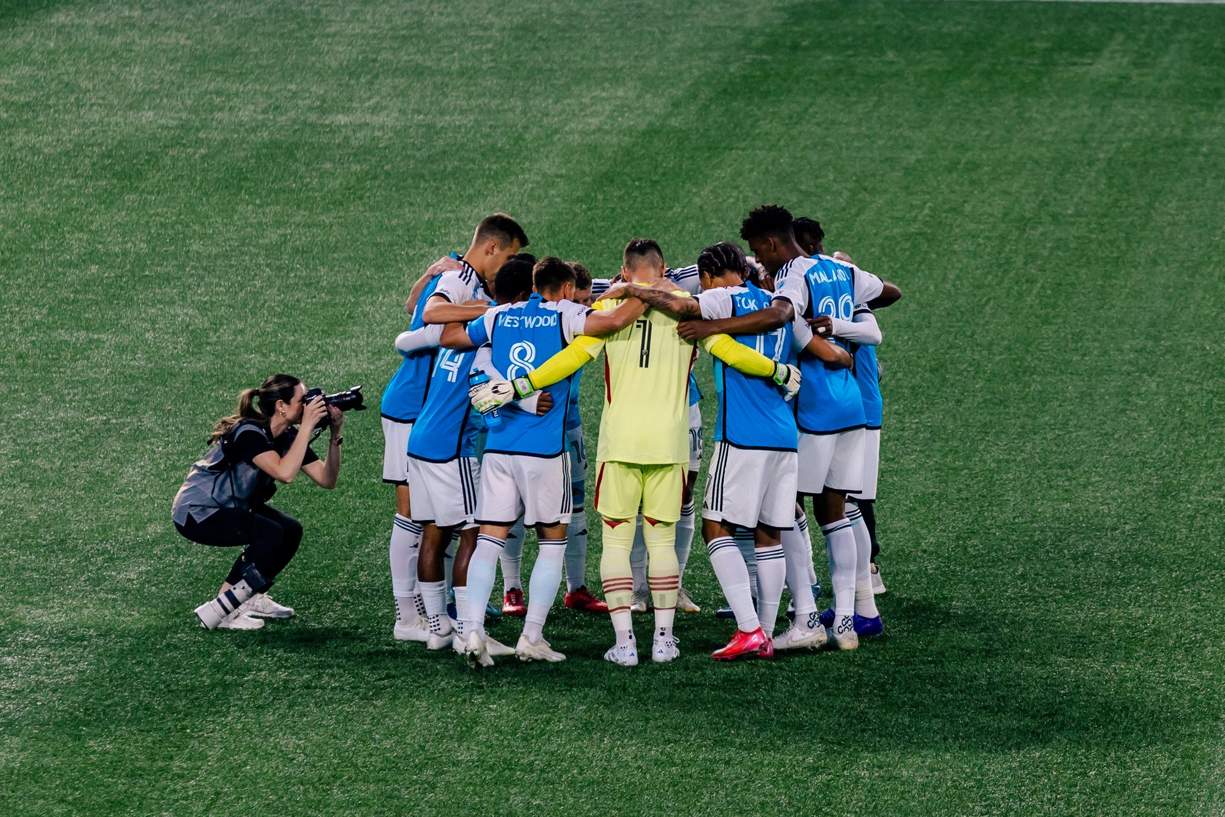 Soccer team huddle on green field in light blue uniforms with photographer capturing the moment.