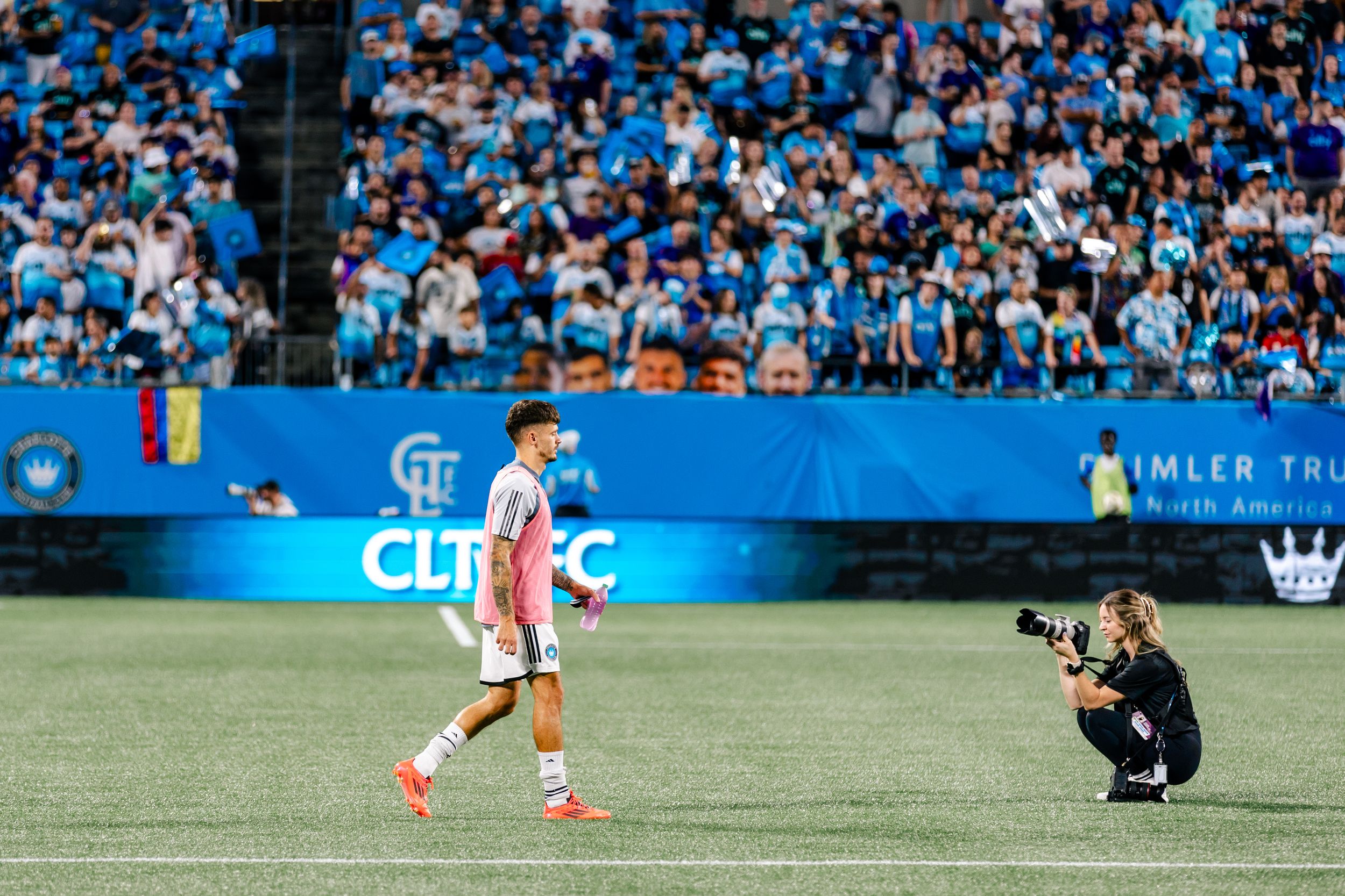 Soccer player in pink uniform walks past crouching photographer on green field.