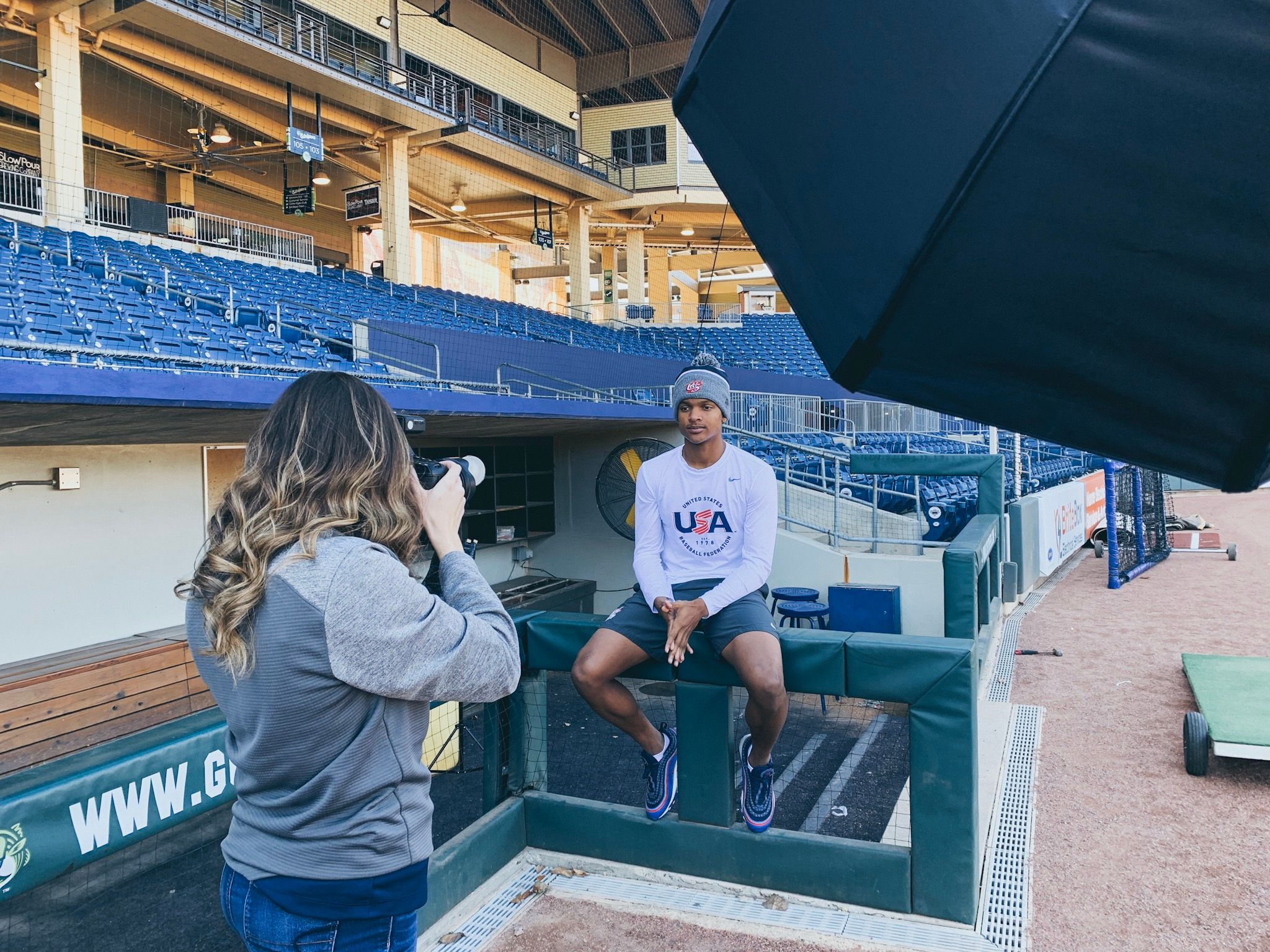 A photographer takes a portrait of an athlete in a USA sweatshirt sitting in a baseball dugout.