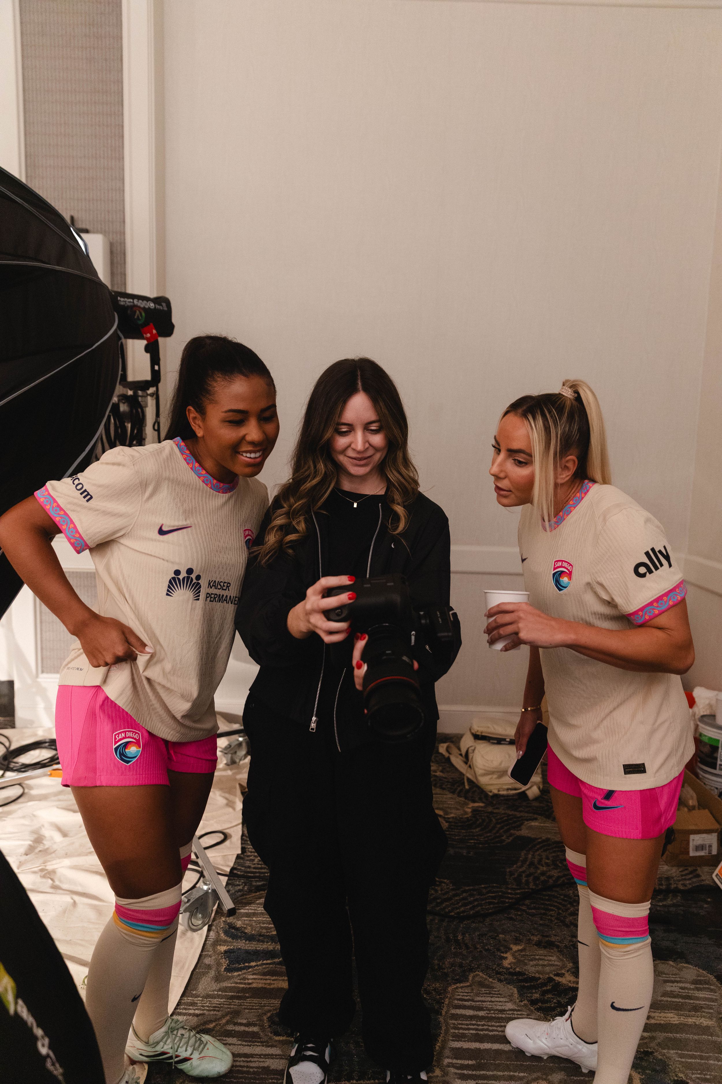 Three women in Nike soccer uniforms with pink shorts review photos on a camera during a team photo shoot.