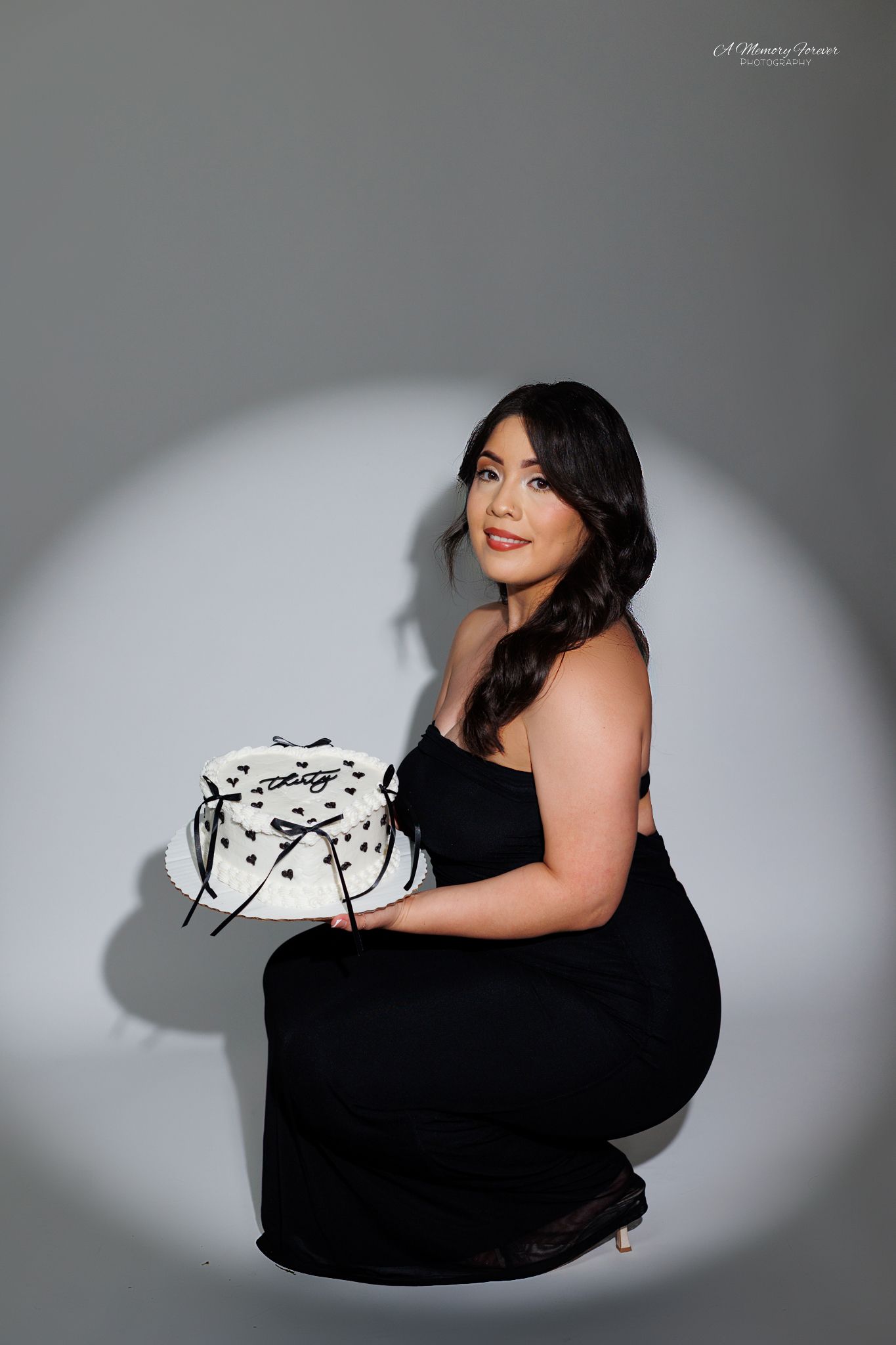 Woman in black dress crouching while holding a decorative white cake in a spotlight photo shoot.