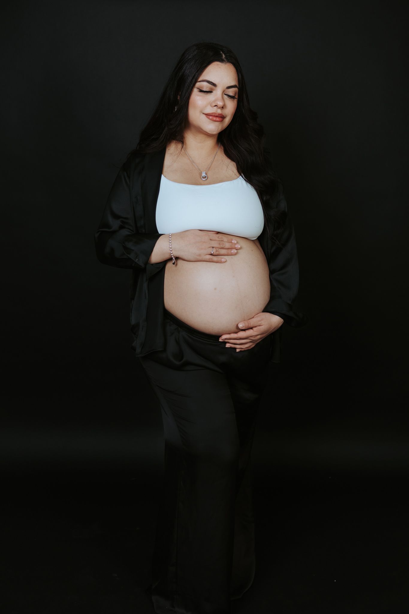 Pregnant woman in white crop top and black outfit cradling her baby bump against a dark studio background.
