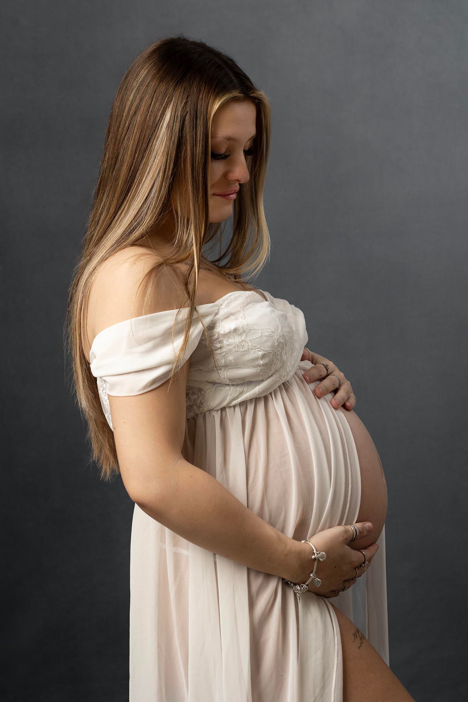 A pregnant woman in a flowing white dress poses for a maternity photo. Tiny moments photography, newborn & maternity photographer in Gosport