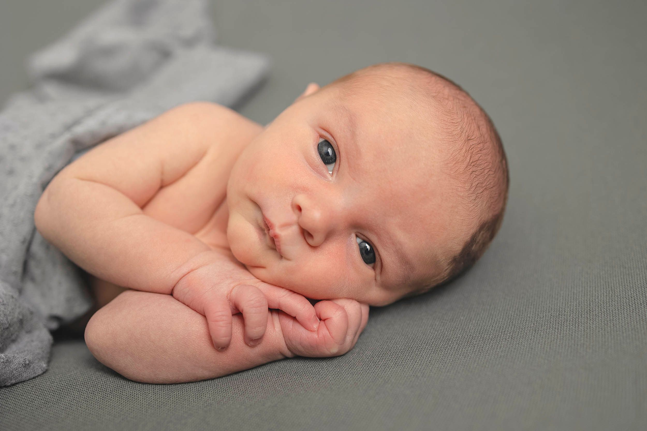 Sweet baby lying on grey blanket looking at camera while gently touching their face. Newborn photography in Gosport and Fareham, Hampshire