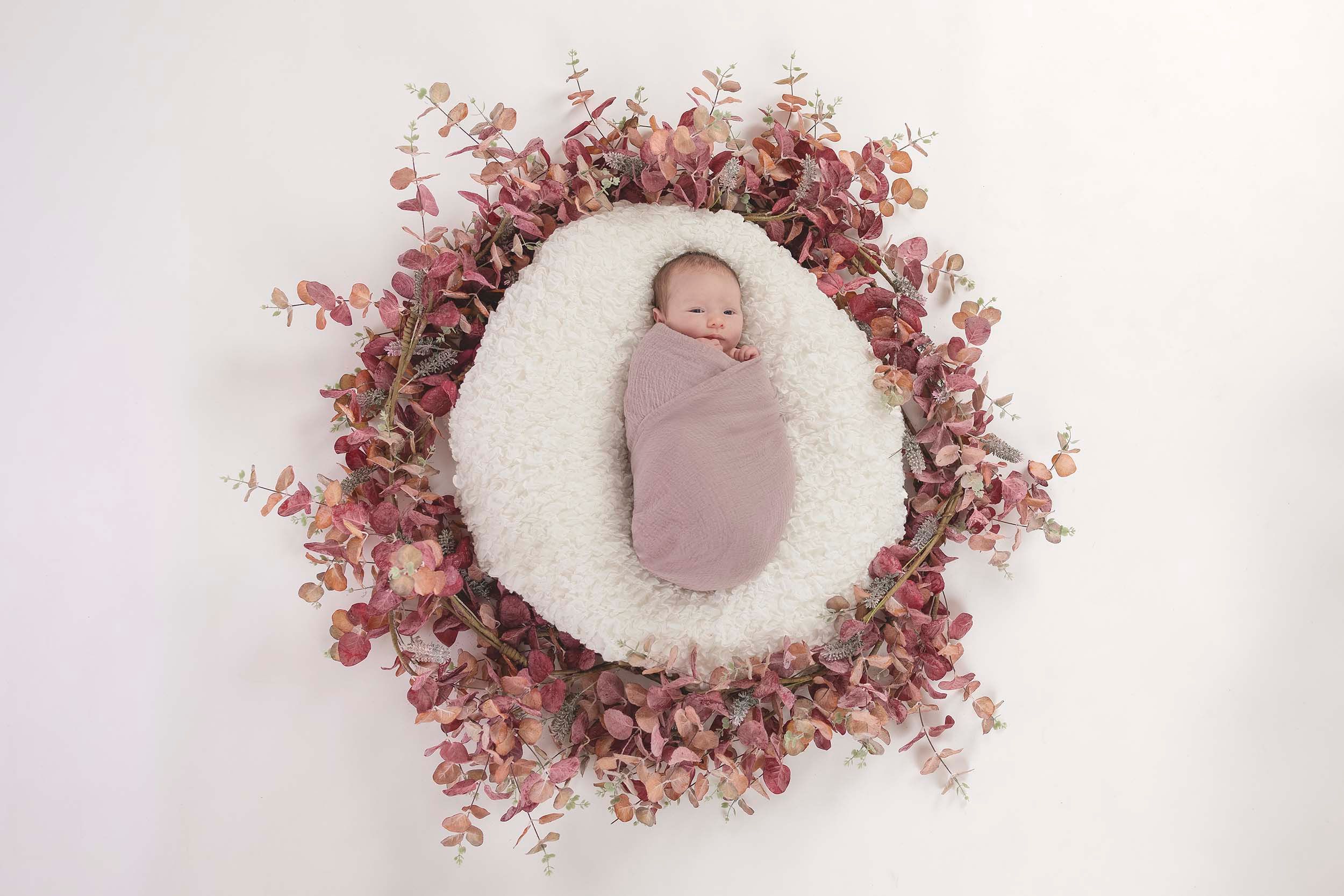 Newborn swaddled in pink fabric surrounded by delicate pink floral decorations. Newborn photography in Gosport, Fareham, Portsmouth