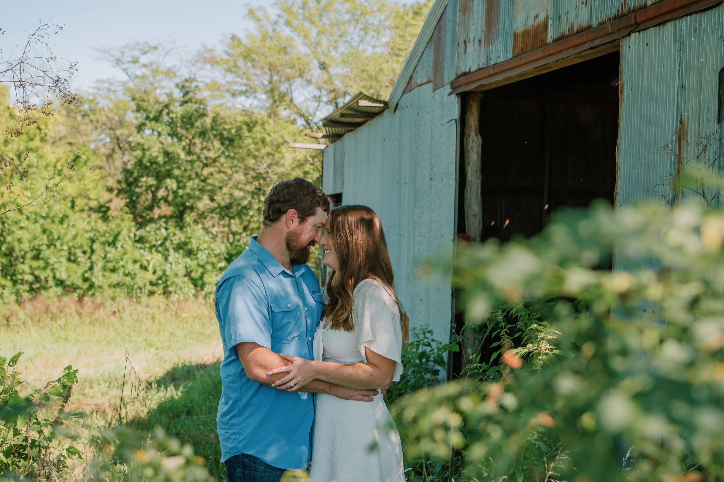 Engaged couple sharing a tender moment in a grassy field under a tree, with a rustic barn in the background, capturing their love