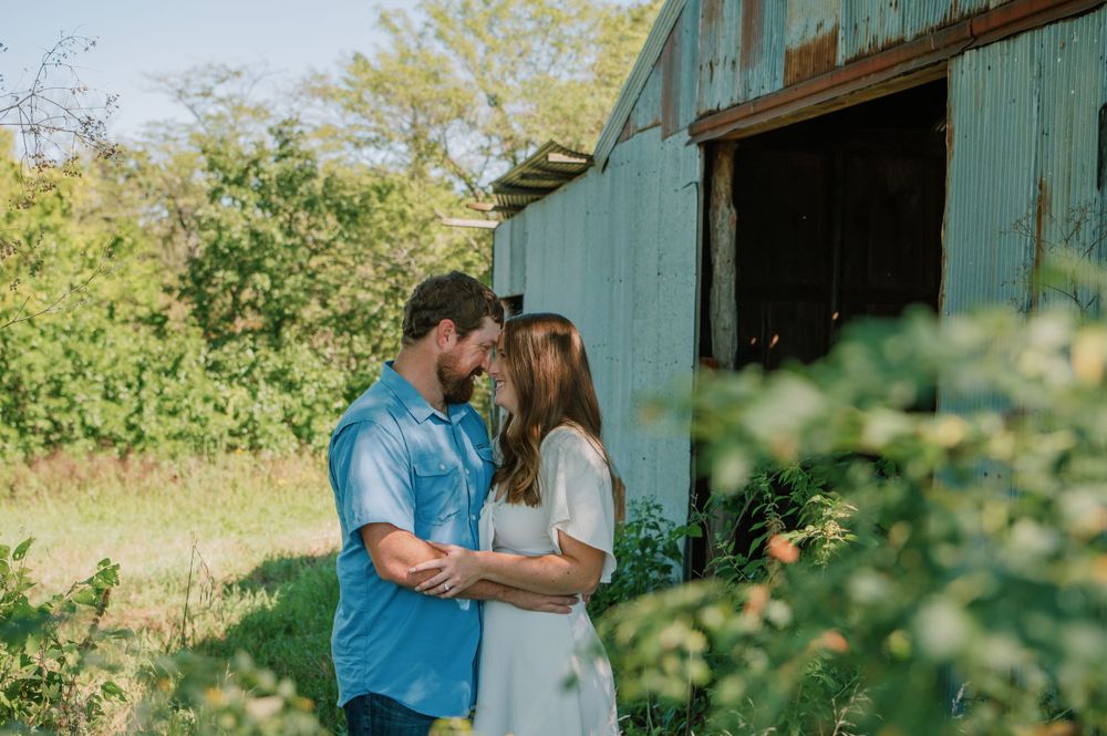 Engaged couple sharing a tender moment in a grassy field under a tree, with a rustic barn in the background, capturing their love