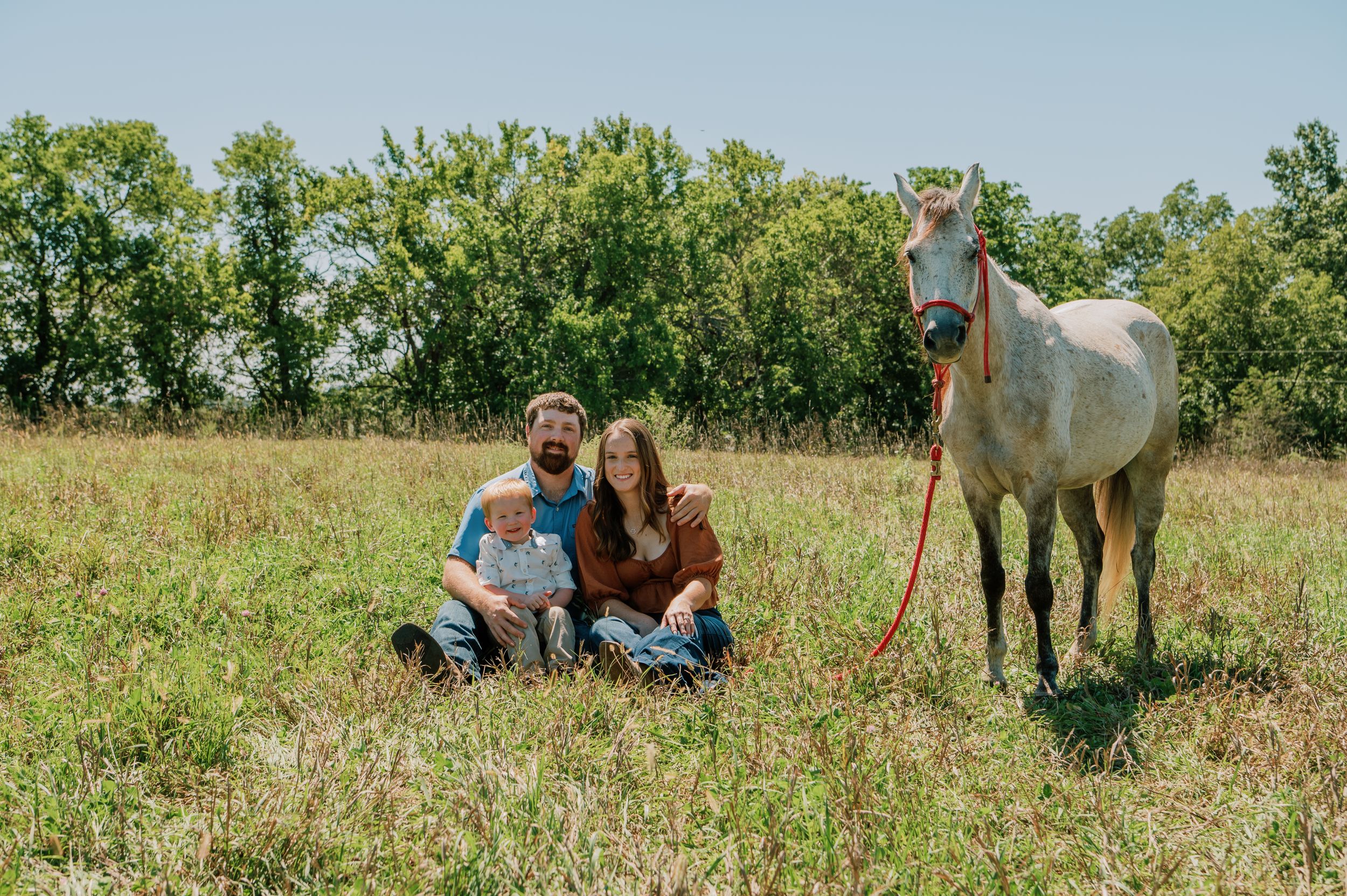 Family of three sitting together in a field, with a horse standing beside them, capturing a peaceful and natural moment