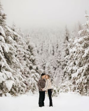 A couple shares a romantic moment surrounded by snow-covered evergreen trees in a misty winter forest.