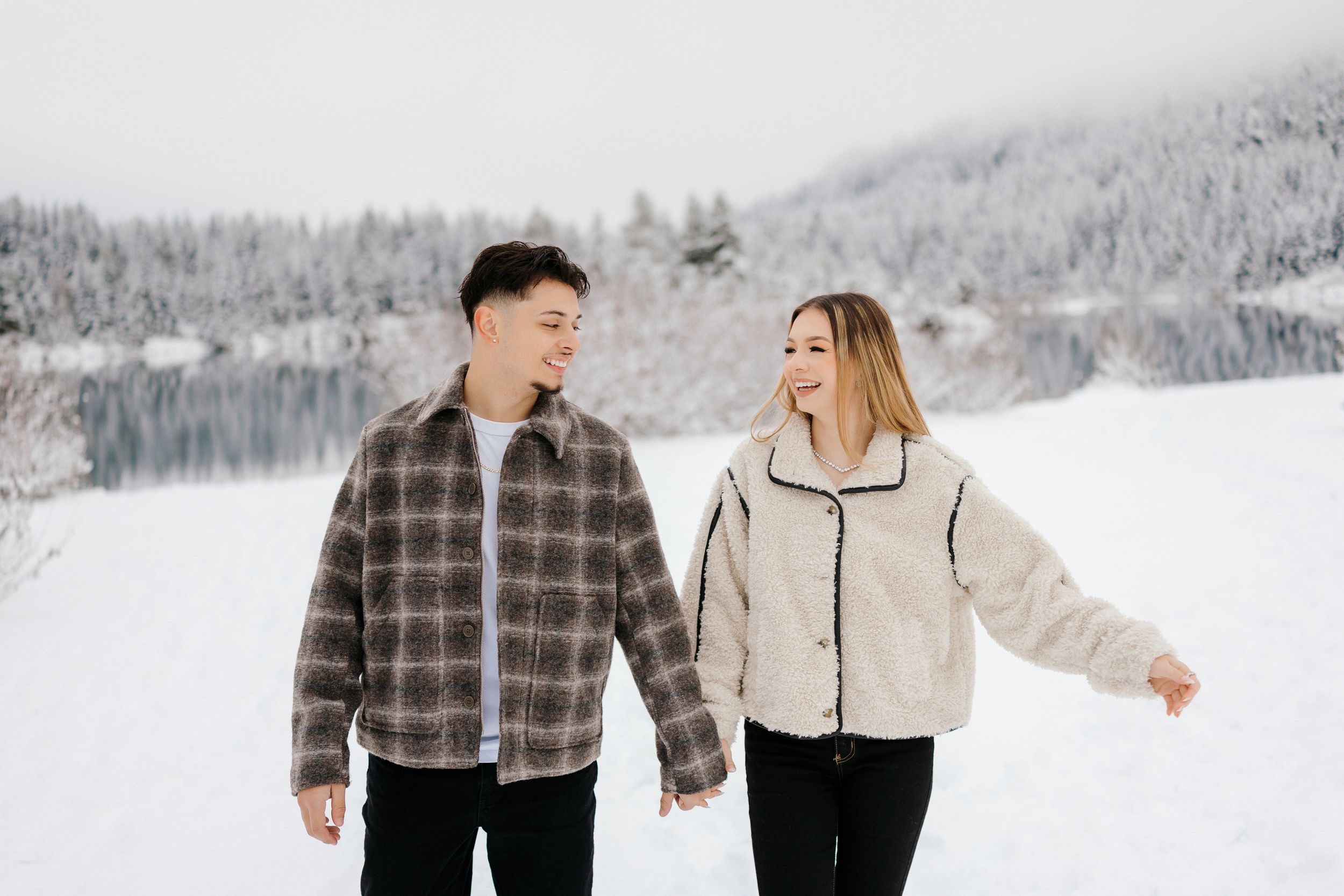 A couple holding hands while walking near a snow-covered lake with snowy mountains and forest in winter.