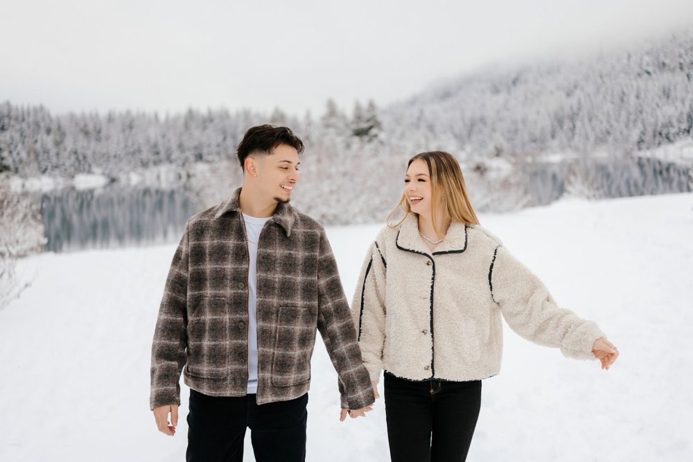 A couple holding hands while walking near a snow-covered lake with snowy mountains and forest in winter.