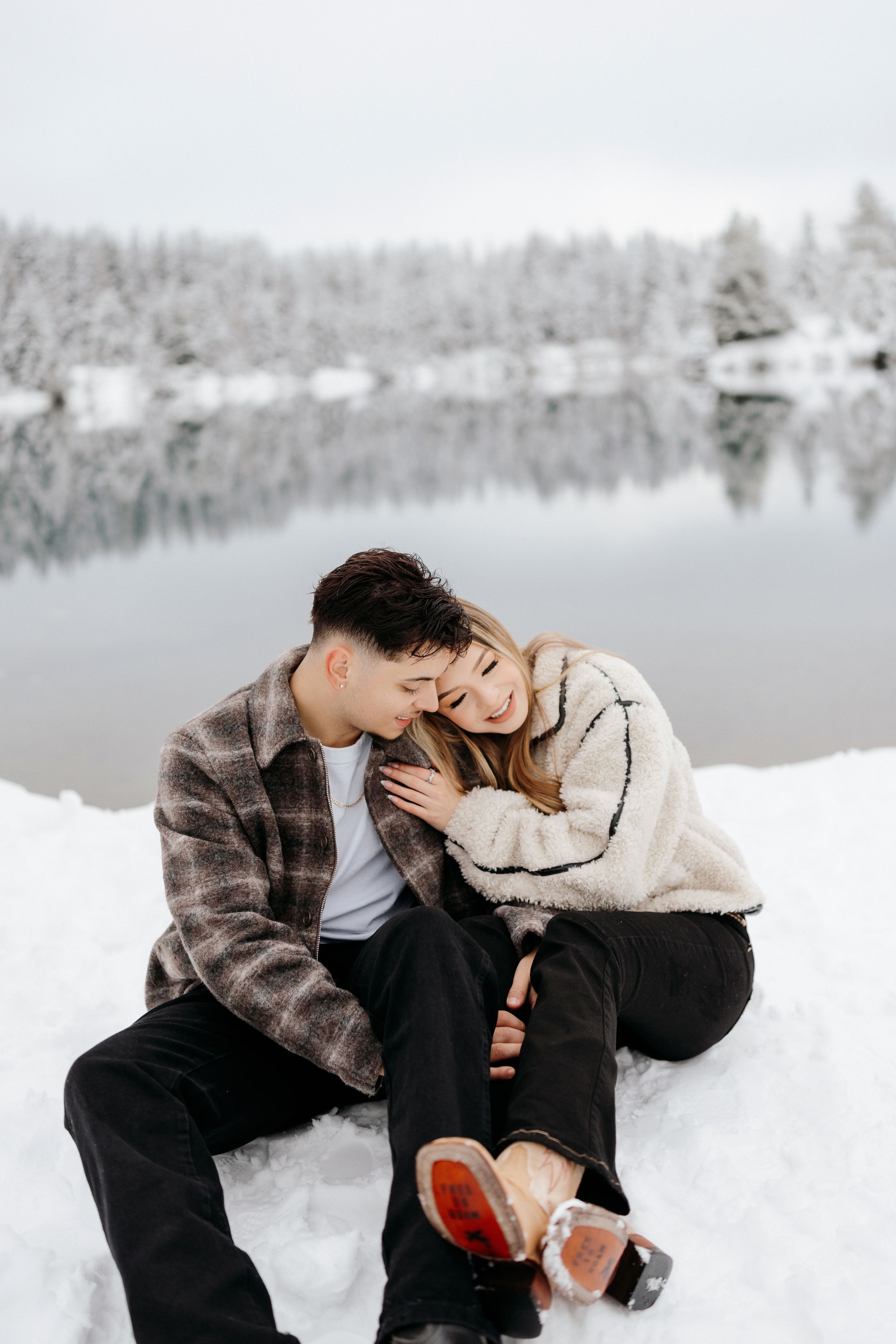 A couple snuggles together while sitting in the snow beside a serene winter lake with snow-covered trees in the background.