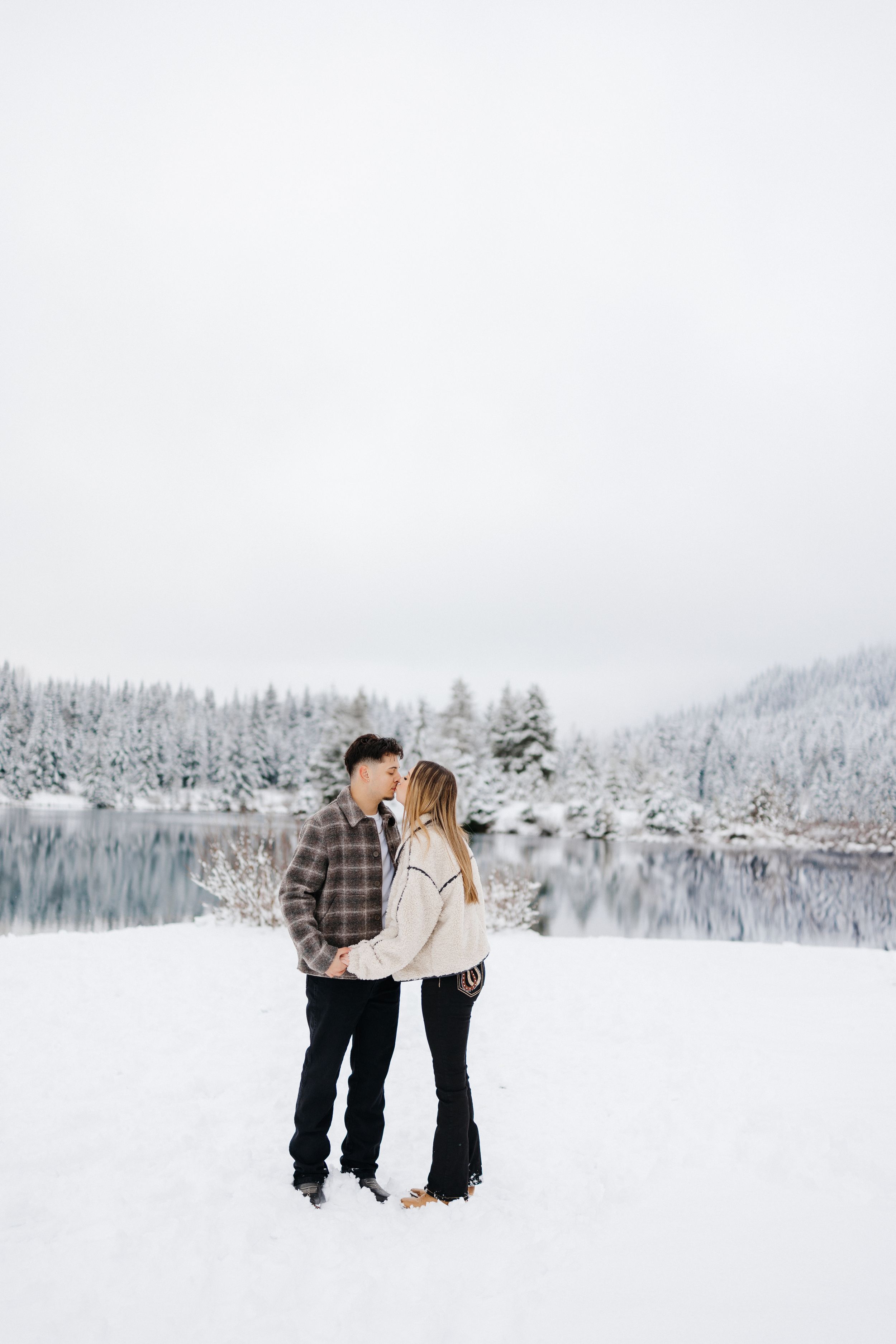 A couple shares a romantic moment on a snowy lakeshore surrounded by snow-covered evergreen trees and mountains.