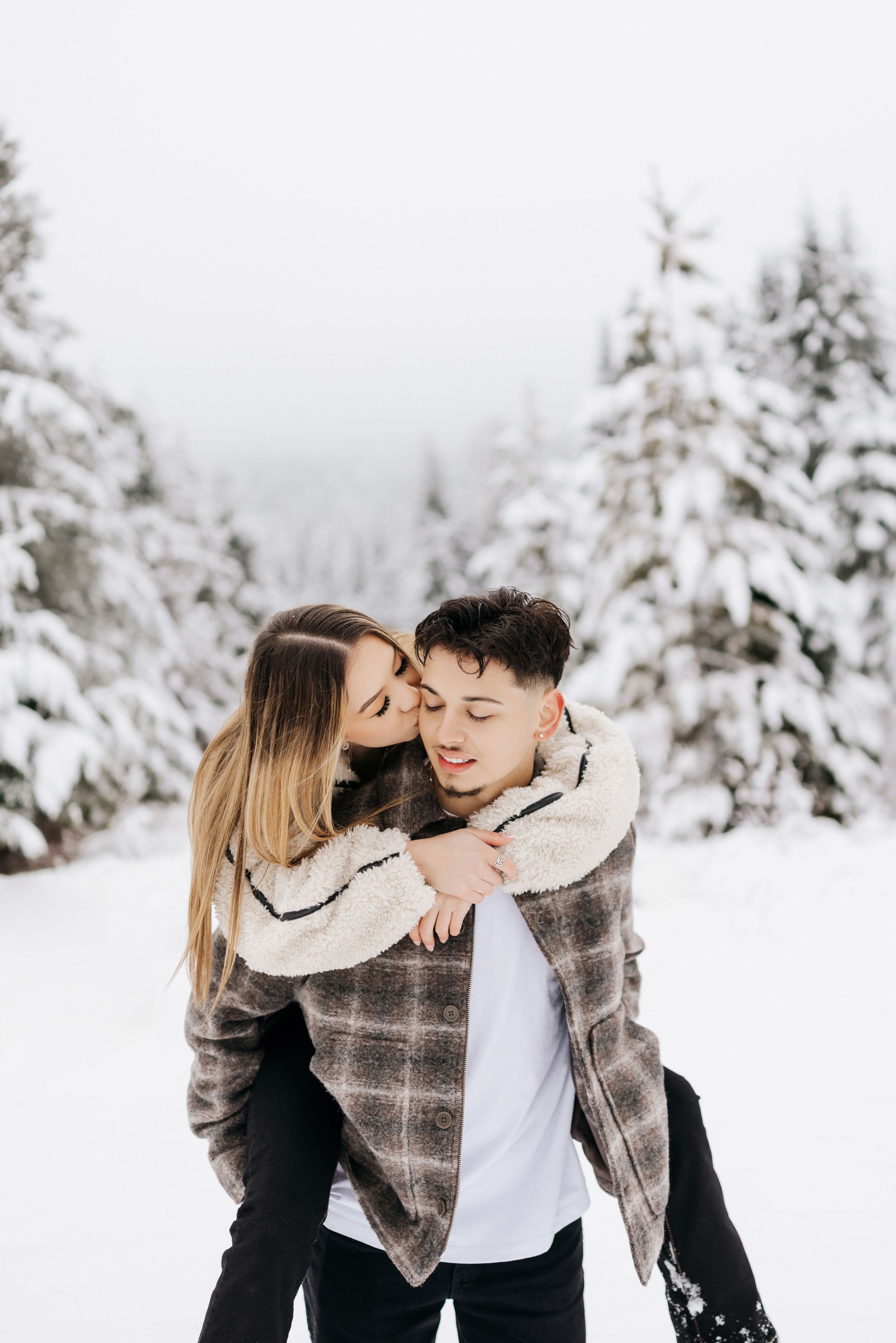 A couple shares an intimate moment while playing in a snow-covered forest landscape during winter.
