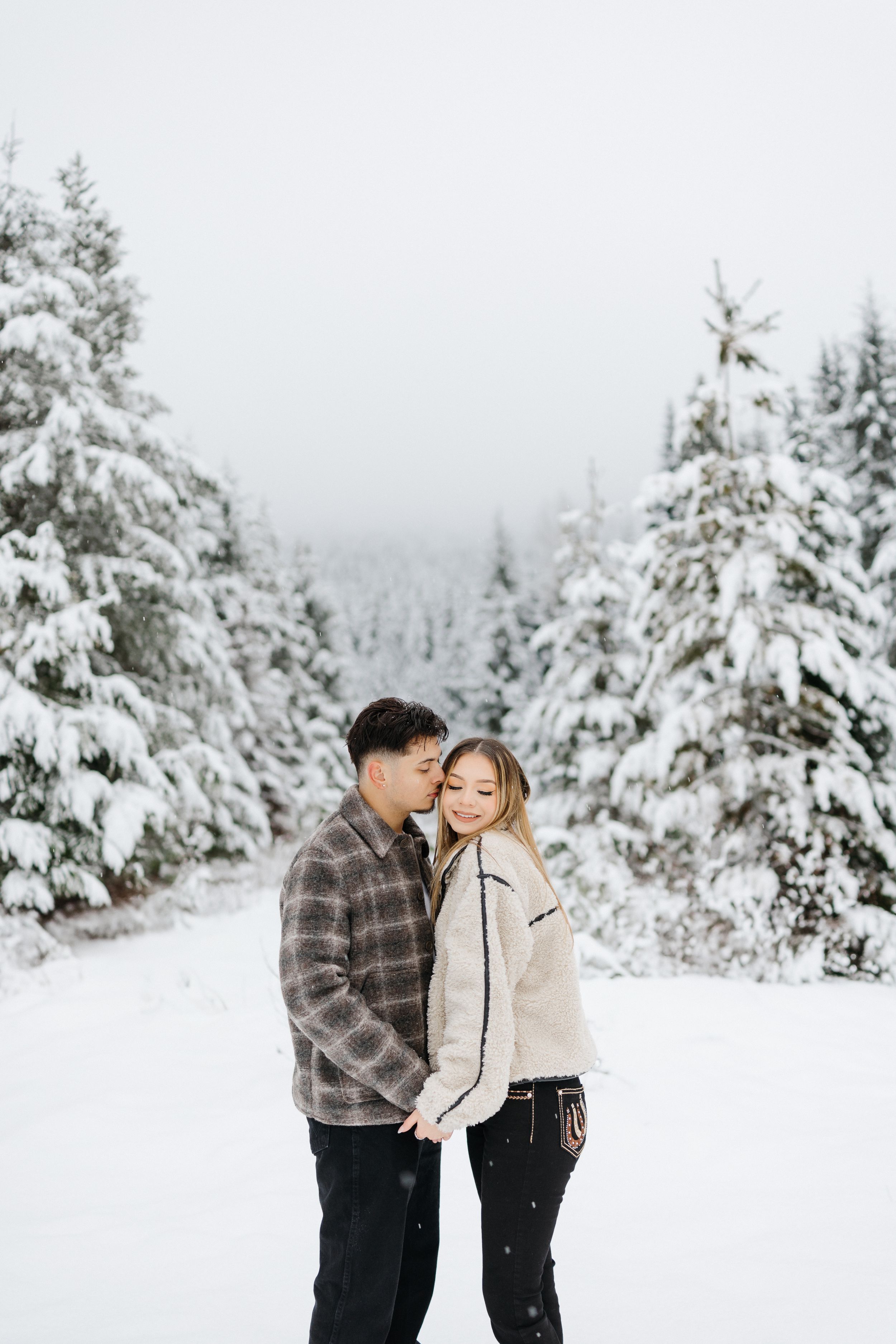 Couple enjoying a romantic winter moment surrounded by snow-covered evergreen trees on a foggy mountain path.
