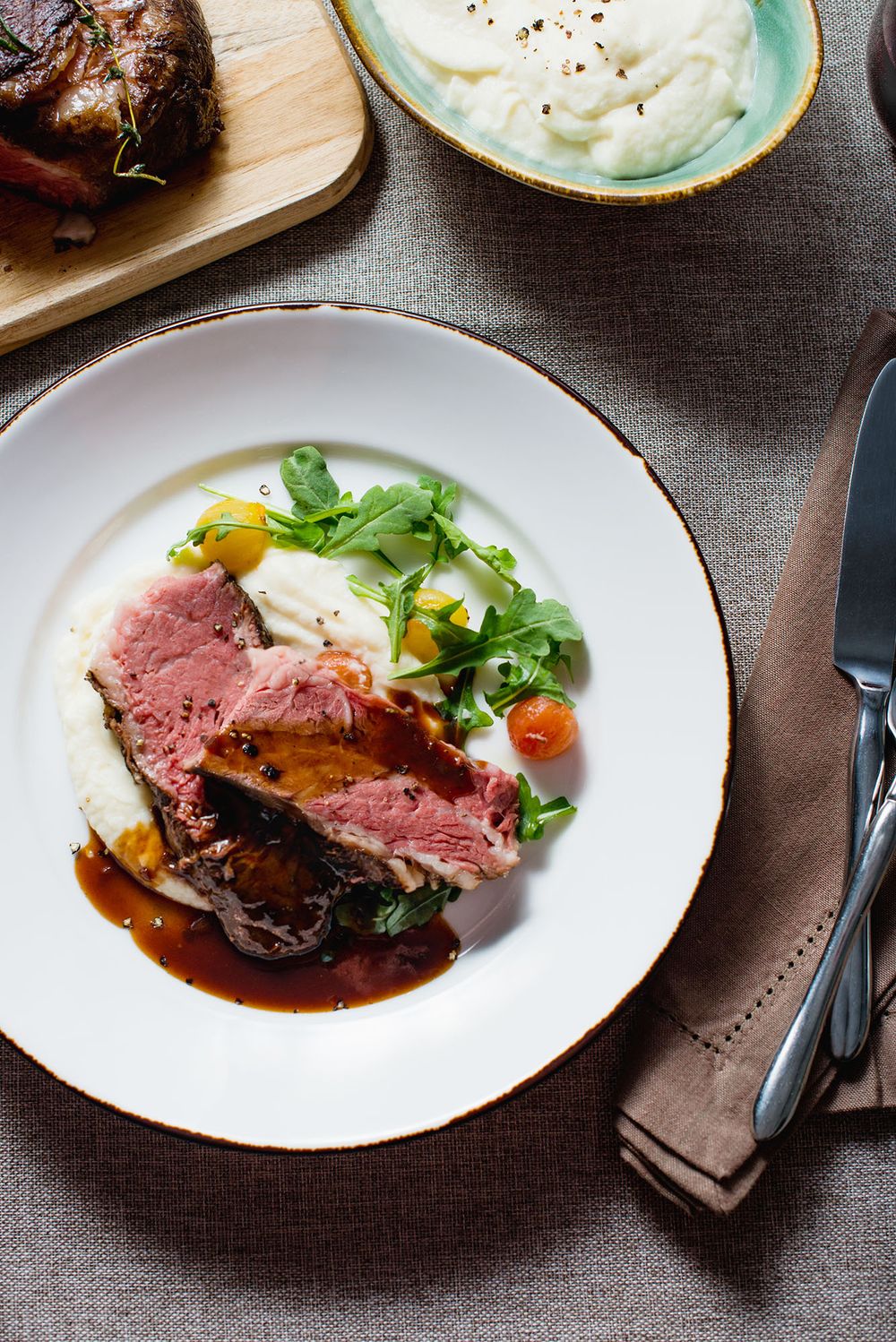 Elegant plated dish with sliced medium-rare beef, mashed potatoes, cherry tomatoes, arugula, and rich brown sauce.