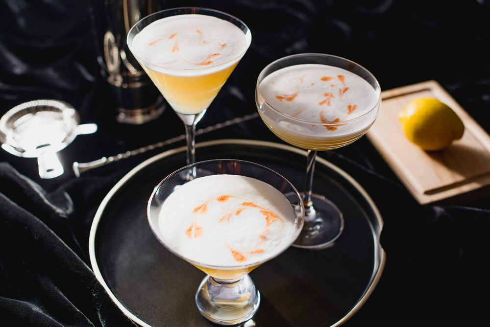 Three elegant cocktails in martini glasses with frothy tops and orange garnish on a dark reflective tray.