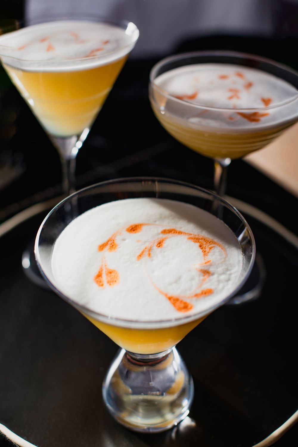 Three elegant cocktails with frothy tops and orange garnish served on a dark round tray.