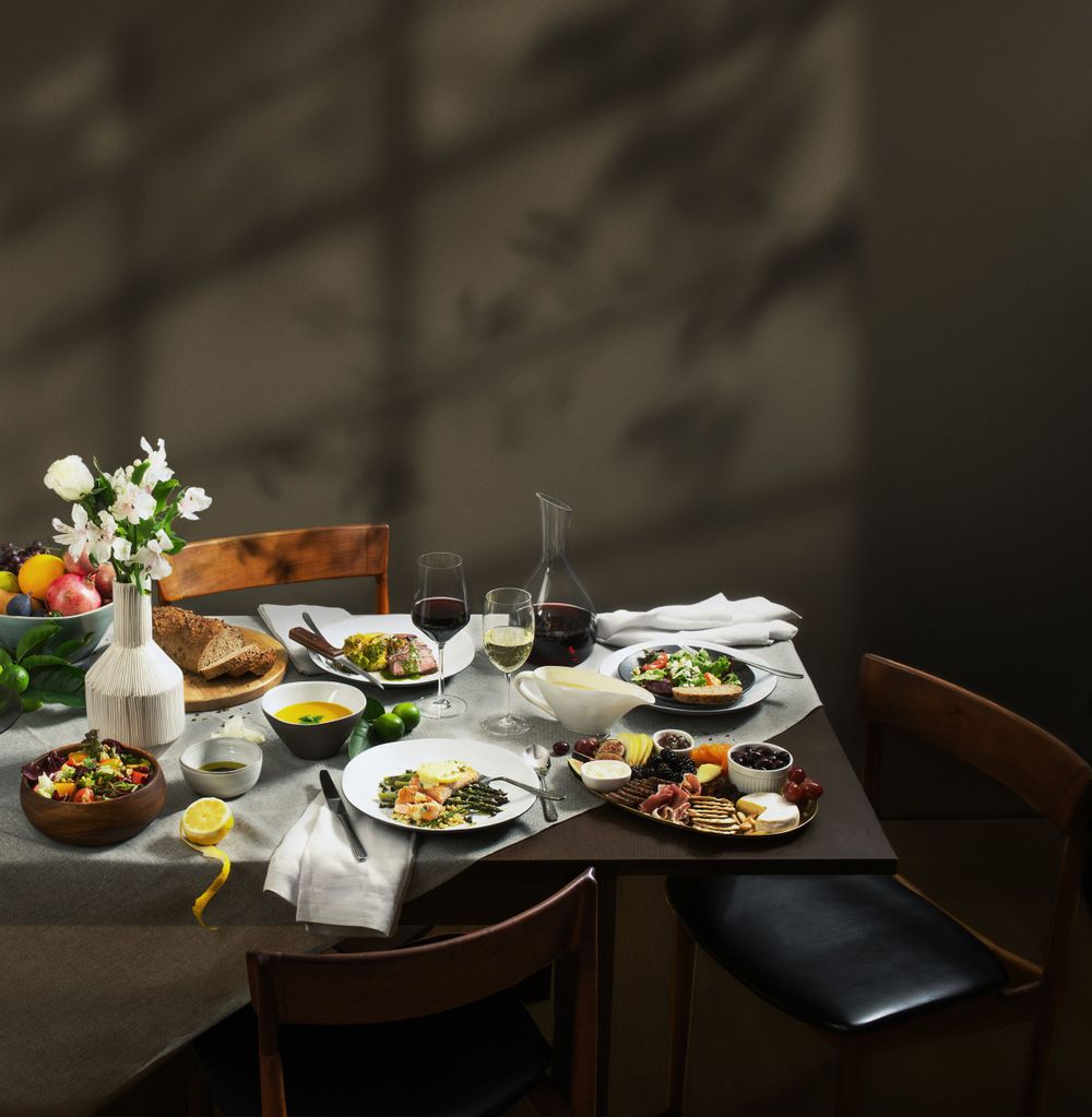 An elegantly set dinner table with multiple dishes, wine glasses, a carafe, and white flowers in soft natural light.