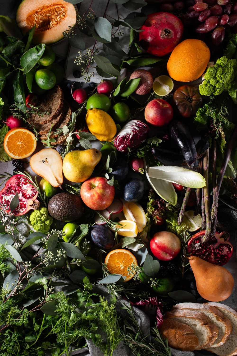 A vibrant overhead flat lay of colorful fresh fruits, vegetables, herbs, and bread arranged on a dark surface.