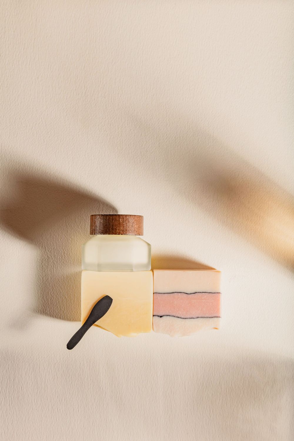 Minimalist skincare product display with glass jar, pink layered soap bar, and spatula.
