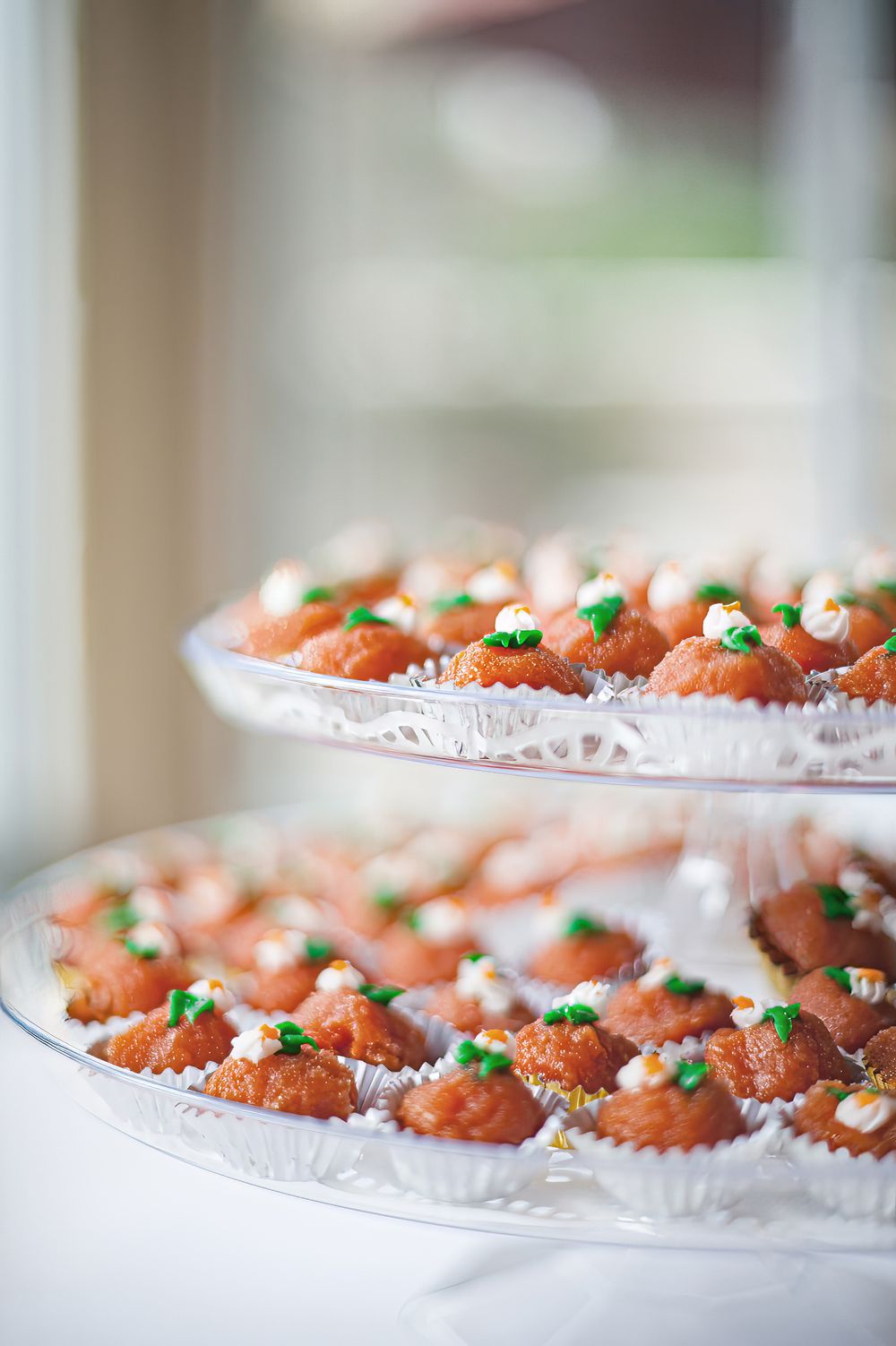 Two-tiered display of mini carrot cake cupcakes with cream cheese frosting and green leaf decorations on white doily plates.